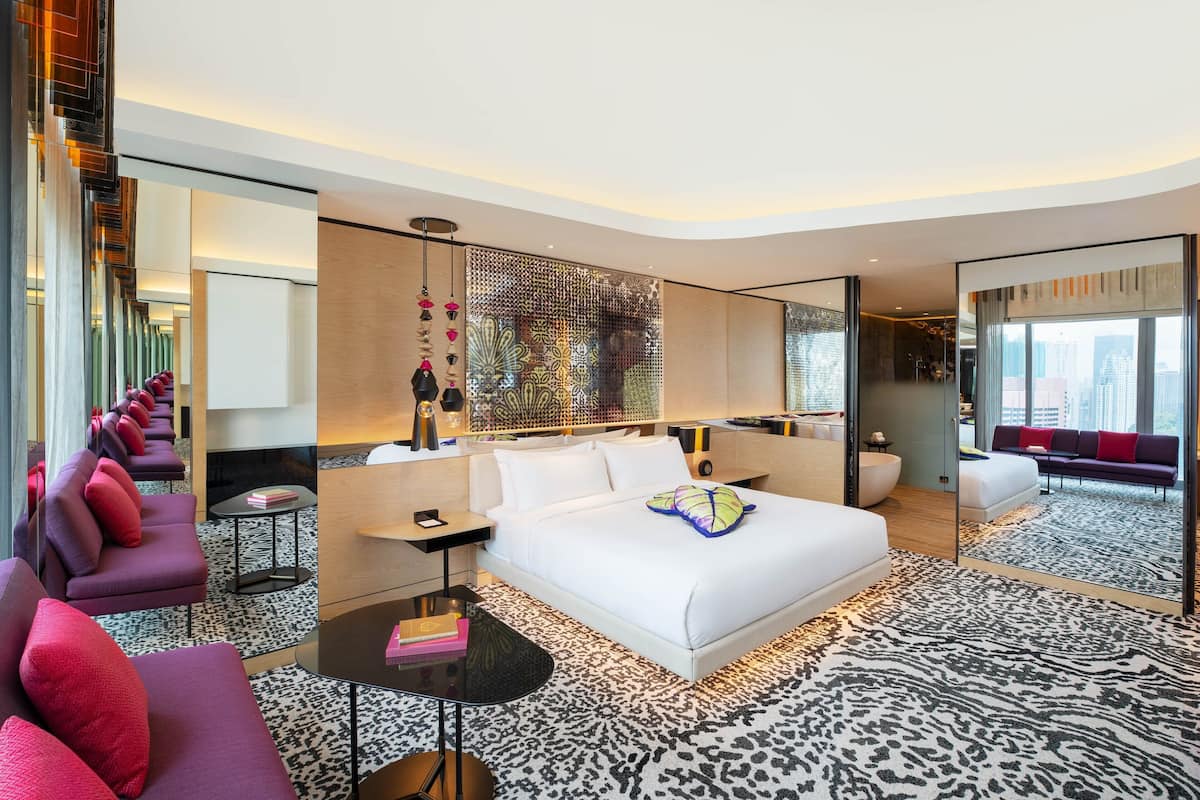 A modern hotel room with a large bed, a seating area with purple sofas, a black coffee table, and a patterned rug.