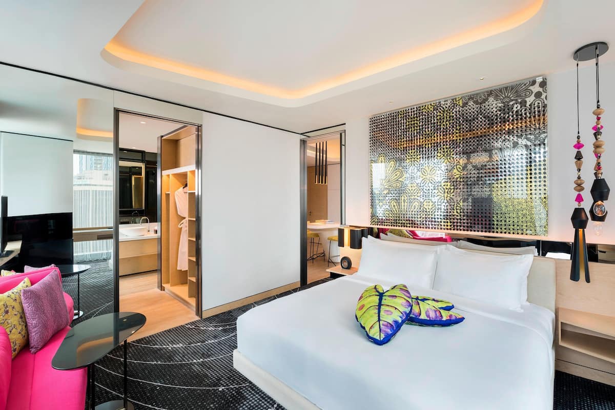 A modern hotel room with a large bed, a desk, a wardrobe, and a bathroom visible through an open door.