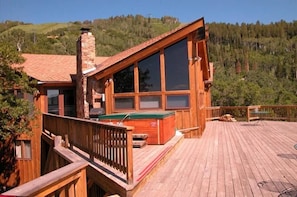 A wooden cabin with large windows, a stone chimney, and a deck with a hot tub.