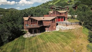 A large, multi-story wooden house with a balcony, set on a hillside with a view of the surrounding landscape.