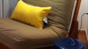 Una cama con un almohadón amarillo, una lámpara de pie y un cubo de basura azul.