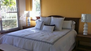 Una cama bien tendida con una colcha estampada y almohadas, una lámpara de noche y una ventana con persianas.