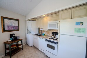 A compact kitchen with white appliances, a microwave, a stove, and a dishwasher. There is a small table with a coffee maker and a framed picture on the wall.