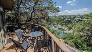 A deck with chairs and a table overlooking a scenic landscape with trees and a body of water. - Cool Treehouse Cabin for wake sport and boating with cave pool [#12 Honeysuckle] (Lakeway)