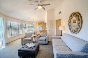 A living room with a sofa, two armchairs, a coffee table, a ceiling fan, a dining area with a table and chairs, and a kitchen area.