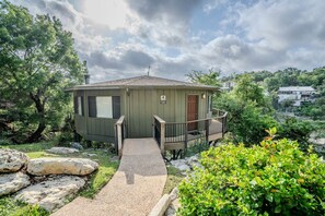 A small, single-story house with a porch and a small deck, surrounded by trees and rocks.