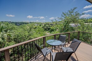 A wooden deck with a glass table and chairs, overlooking a lush green landscape with trees and houses.