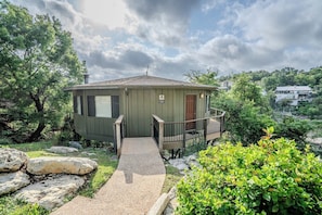 A small, single-story house with a porch and a small deck, surrounded by trees and rocks.