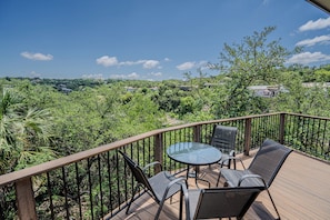 A deck with a glass table and chairs, surrounded by a wooden railing, overlooking a lush landscape with trees and houses.