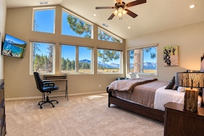 A bedroom with a bed, desk, and chair, large windows with mountain views, and a ceiling fan.