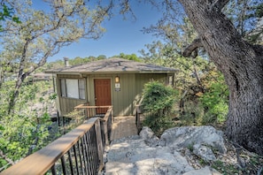 A small, single-story cabin with a wooden deck and a front door, surrounded by trees and natural scenery. - Engelman`s Daisy #11 - Elevated Treehouse with Pool & Hot Tub Access (Lakeway)