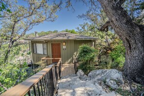 A small cabin with a wooden deck and a door, surrounded by trees and rocks.