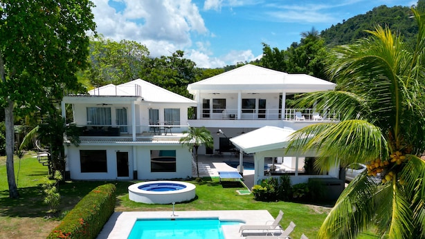 Une maison blanche à deux étages, dotée d’une piscine, d’un espace salon extérieur et d’une végétation luxuriante.