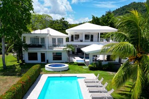 Casa blanca de dos pisos con piscina, mobiliario exterior y abundante vegetación.