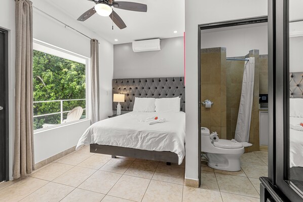Un dormitorio moderno con cama, ventilador de techo, ventana con cortinas y baño con inodoro y ducha.