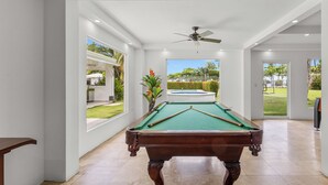 Une table de billard dans une pièce spacieuse, dotée de grandes fenêtres et d’un ventilateur de plafond.