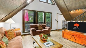 A spacious loft-style room with a high ceiling, large windows, a cozy brown sofa, a wooden coffee table with a basket, and a television on a wooden stand.