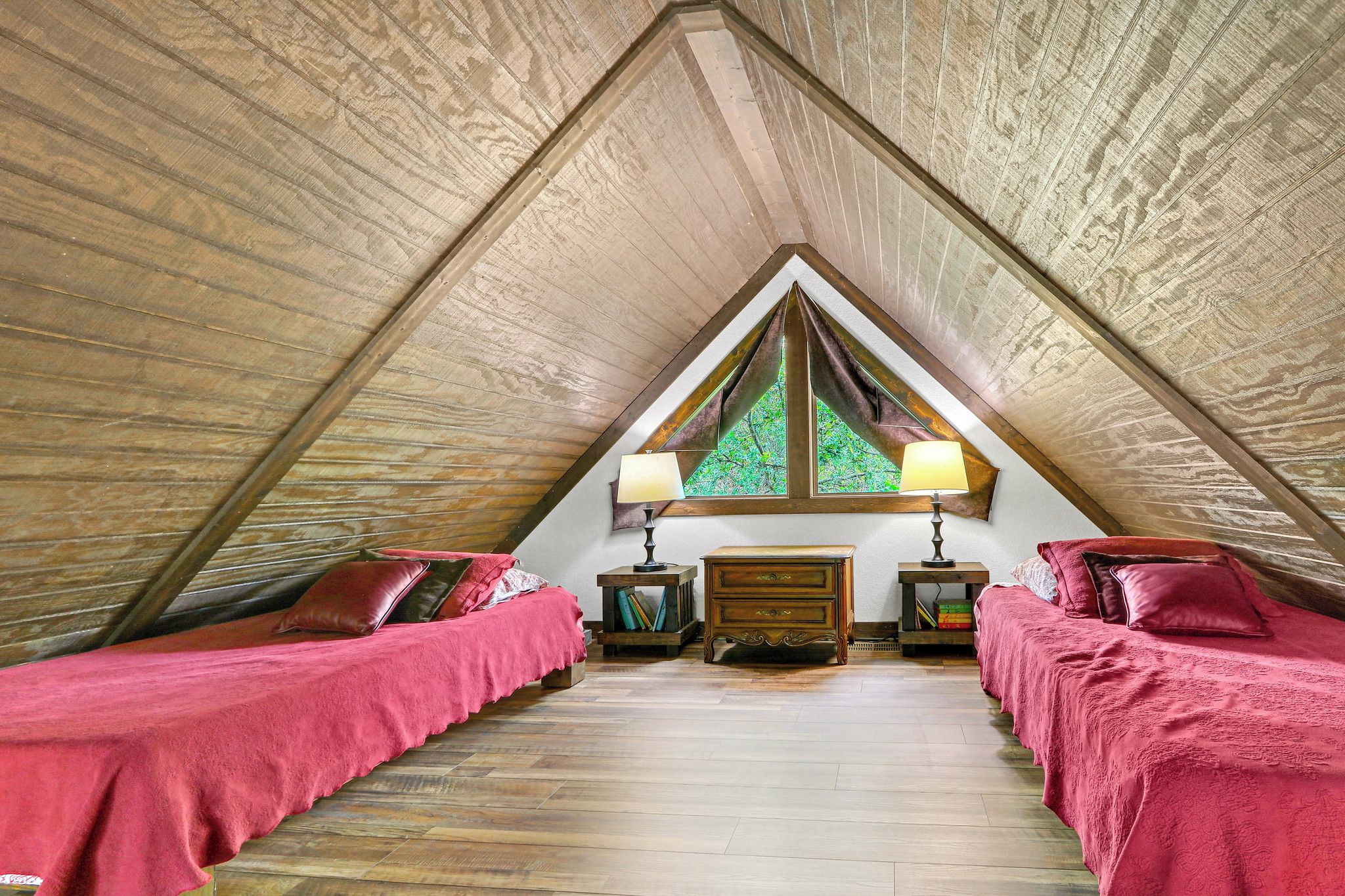 A room with a sloped ceiling, two beds with red bedding, a wooden dresser, two bedside lamps, and a window with a view of greenery.