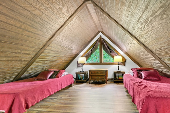 A room with a sloped ceiling, two beds with red bedding, a wooden dresser, two bedside lamps, and a window with a view of greenery.