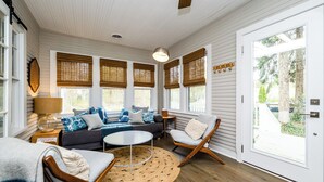 Une pièce chaleureuse avec un canapé, des fauteuils, une petite table et une lampe. De grandes fenêtres sont équipées de stores, et la pièce comprend également un ventilateur de plafond et une porte donnant sur l’extérieur.