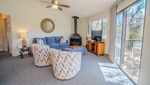 Ein Wohnzimmer mit einer Couch, Fußhockern, einem Kamin, einem Fernseher und einem Balkon mit Gartenmöbeln.