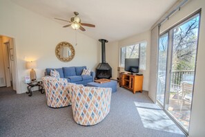 A living room with a sofa, ottomans, a fireplace, a television, and a balcony with outdoor furniture. - Midweek waterfront heaven near Austin with pool and hot tub [#5 Rain Lily] (Lakeway)