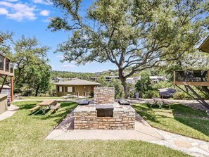 A backyard with a stone fire pit, picnic tables, and a tree providing shade.