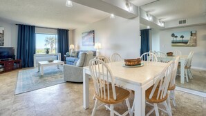 Un coin repas avec une grande table, des chaises blanches et un ensemble de vaisselle bleu. Le salon comprend un canapé et un téléviseur.