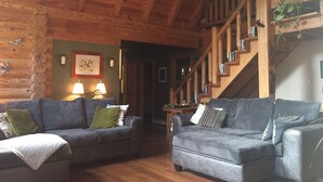 A cozy log cabin interior with two grey sofas, wooden flooring, a staircase, and a green wall with framed artwork. - perfect getaway near sylvan beach! 2 night minimum (Blossvale)