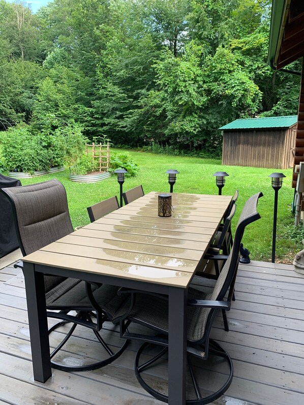 An outdoor deck with a wooden table and chairs, surrounded by a lush garden and a wooden shed. - perfect getaway near sylvan beach! 2 night minimum (Blossvale)