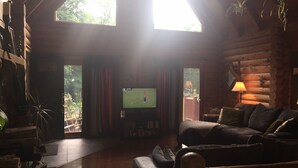 A cozy log cabin interior with a wooden floor, a sofa, a television, and a window overlooking greenery. - perfect getaway near sylvan beach! 2 night minimum (Blossvale)