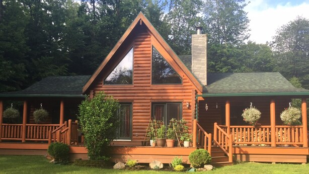 A log cabin with a steeply pitched roof, a chimney, and a porch with hanging flower baskets. - perfect getaway near sylvan beach! 2 night minimum (Blossvale)
