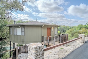 Une petite cabane à un seul étage, dotée d’une terrasse en bois et de murs en pierre, située dans une zone boisée sous un ciel dégagé.