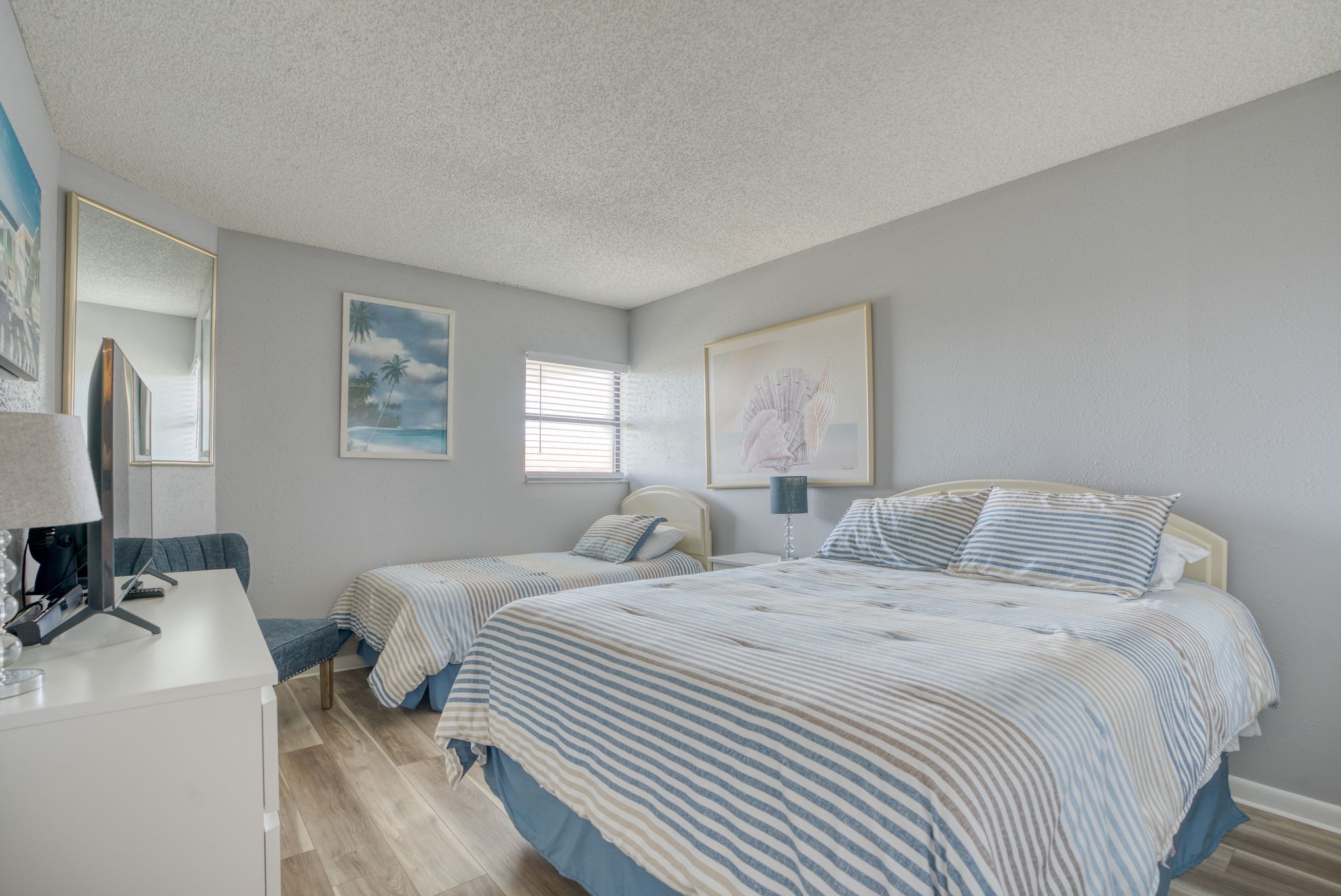 Une chambre avec deux lits simples, un bureau et une chaise. Les murs sont ornés de tableaux encadrés et il y a une fenêtre avec des stores.