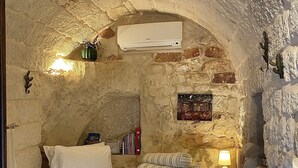 A stone-walled bedroom with a bed, a small table with a lamp, and a wall-mounted air conditioner. - AMMARE BY THE SEA         (Marina di Novaglie)