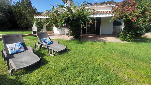 Un patio con dos sillones de descanso y una mesa, rodeado de un césped cuidado y un edificio con techo de tejas.