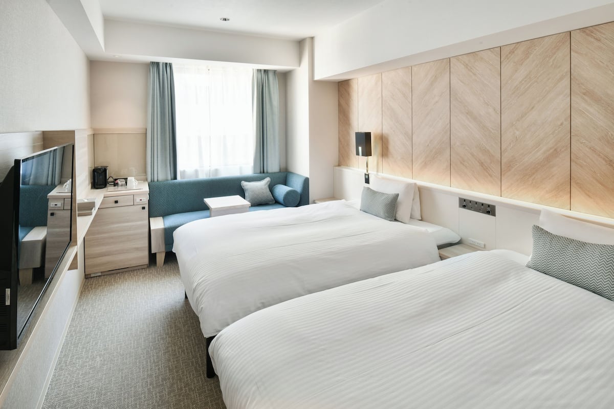 A hotel room with two beds, a desk with a chair, a TV, and a large window with curtains.