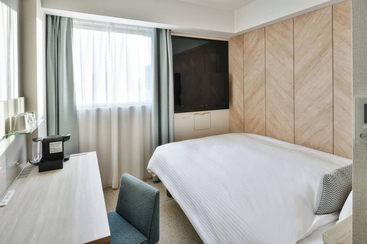 A hotel room with a bed, a desk with a coffee maker, a TV, and a window with curtains.