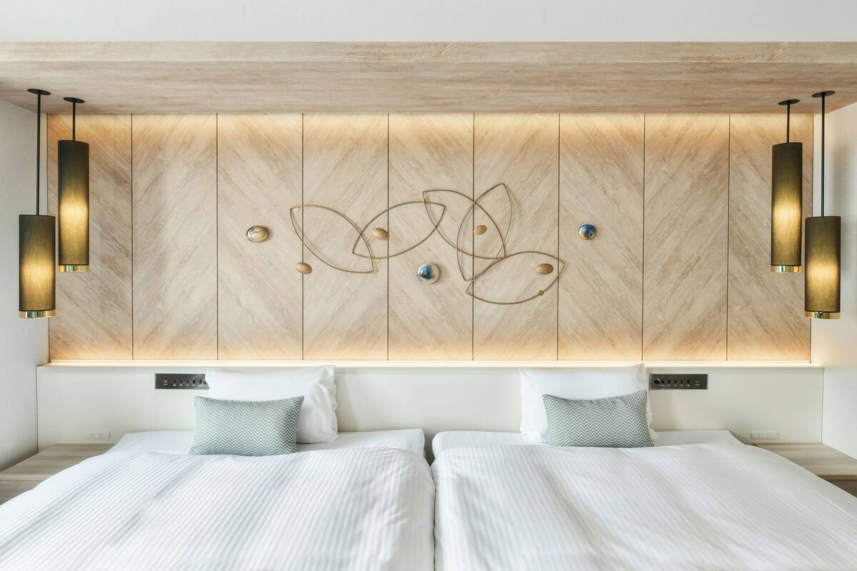 A modern hotel room with a wooden headboard, two beds with white linens, and pendant lighting.