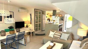Una sala moderna con un sofá, una mesa de centro y sillas. Se ve un televisor, un comedor y una cocina al fondo.