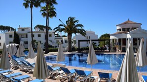 Un área junto a la piscina con sillones de descanso y sombrillas, una piscina azul cristalina y edificios al fondo.