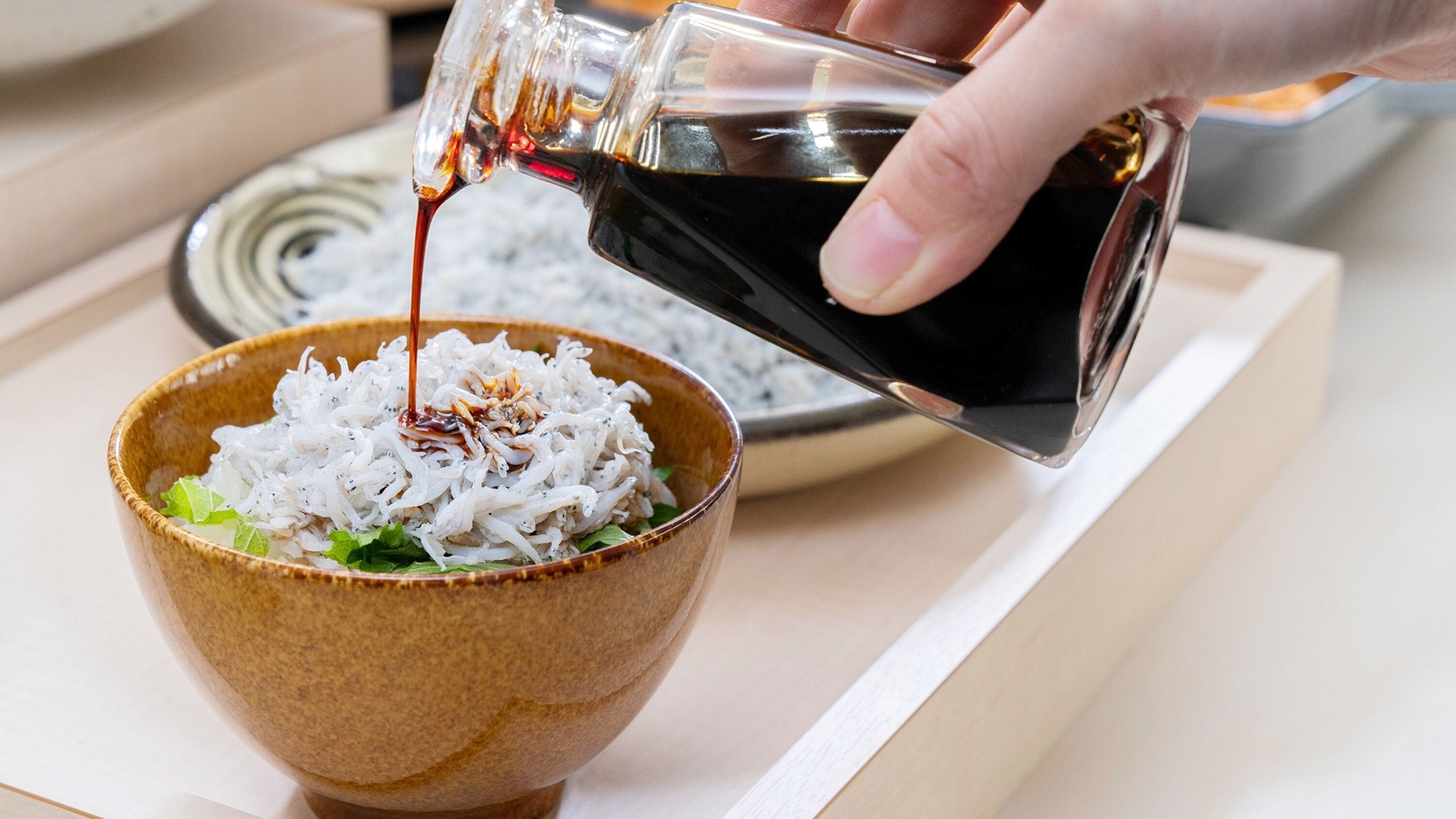 A hand pouring soy sauce over a bowl of rice with vegetables.