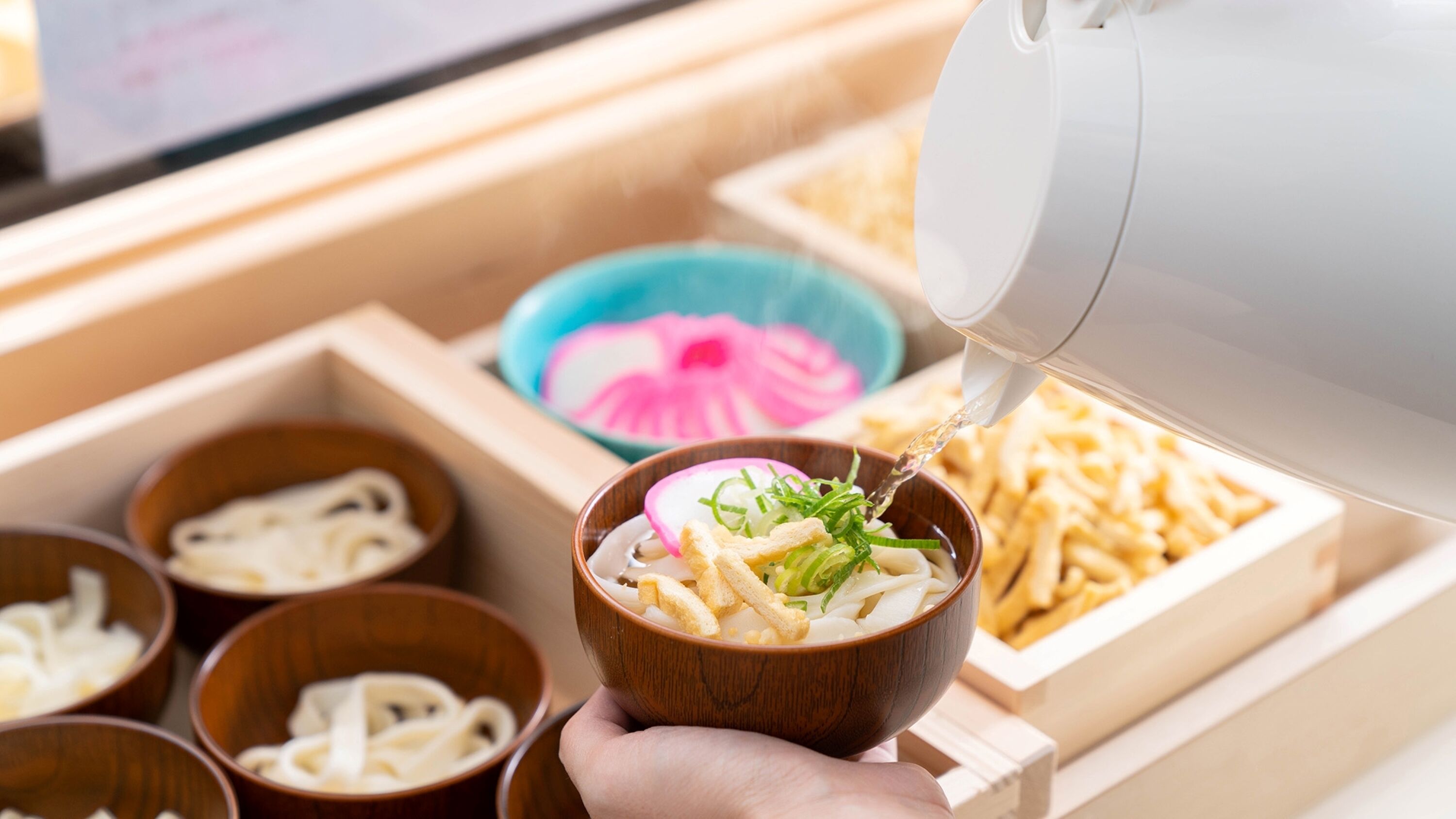 A person pouring a clear liquid into a wooden bowl containing noodles and vegetables.