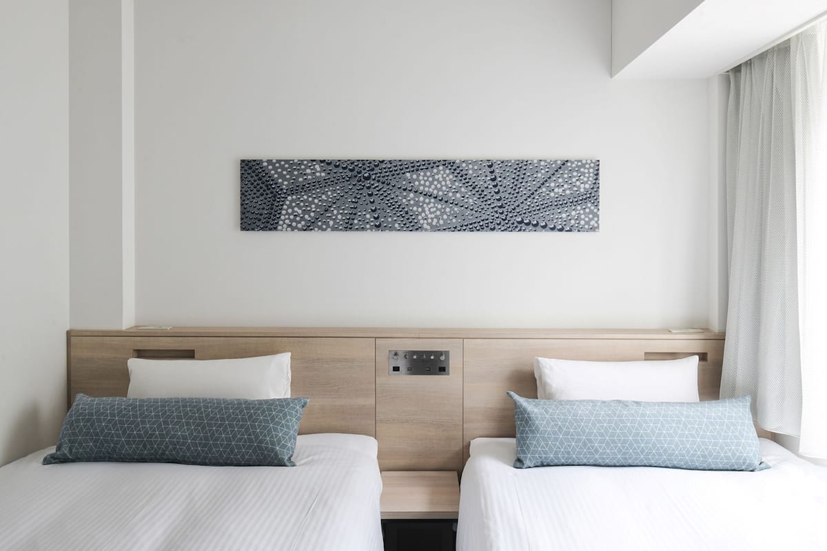 A hotel room with two beds, a headboard with a control panel, and a wall-mounted abstract art piece.