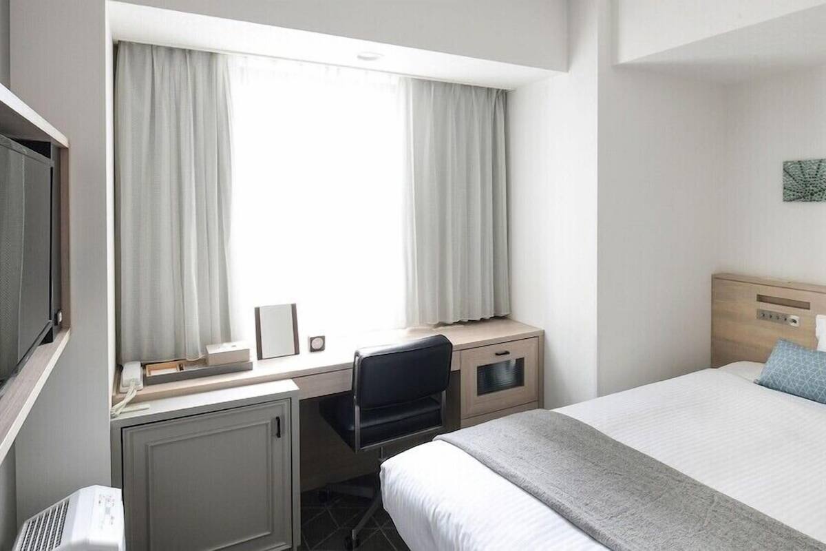 A hotel room with a bed, a desk, a chair, a TV, and a window with curtains.