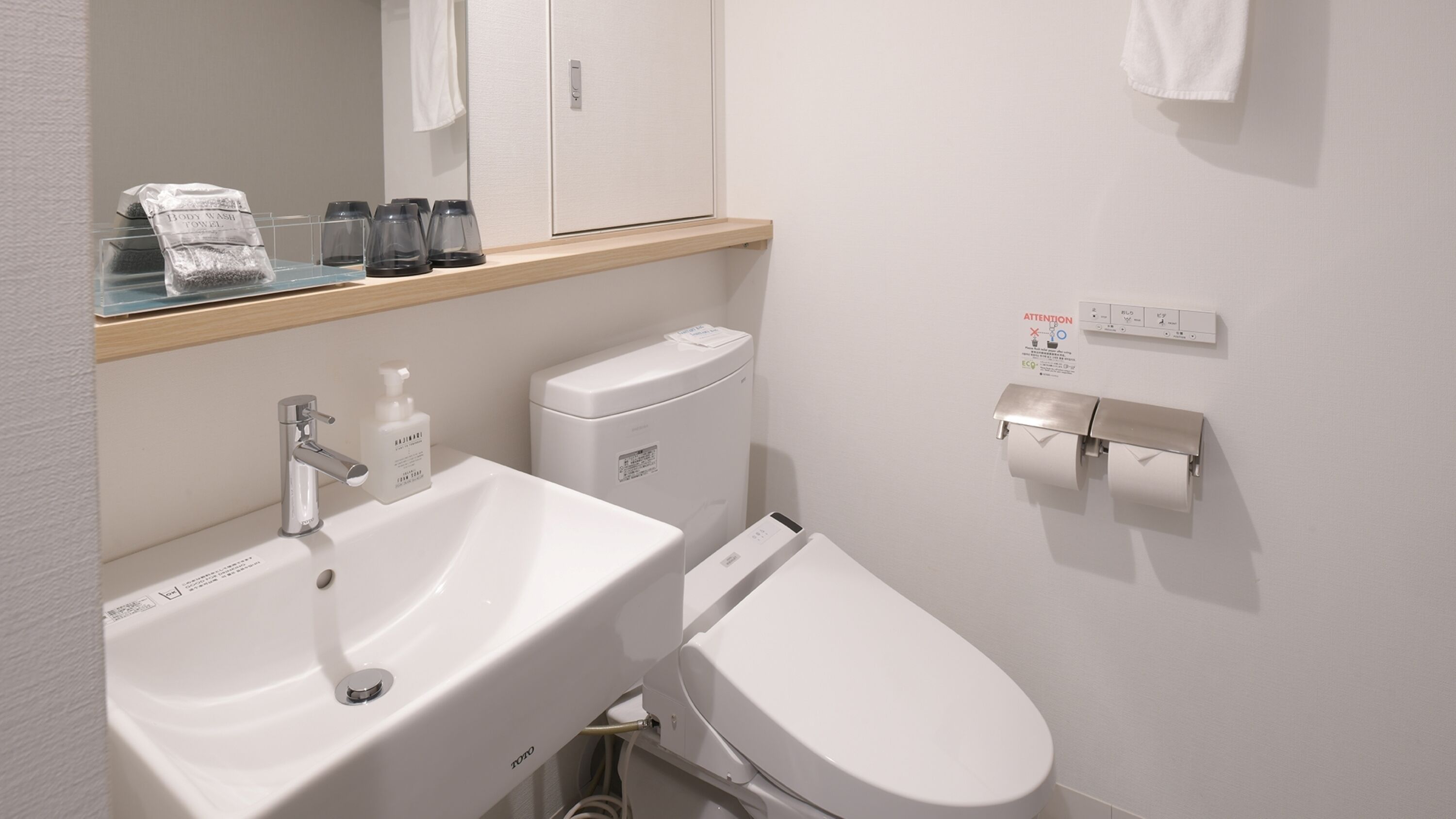 A modern bathroom with a white sink, toilet, and bidet. There is a mirror above the sink, a towel hanging on the wall, and a soap dispenser on the sink.