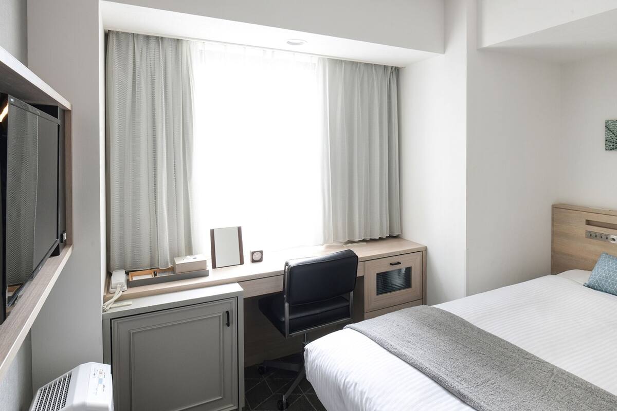 A hotel room with a bed, a desk with a chair, a TV, and a window with curtains.