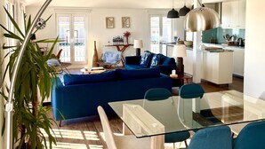A modern living room with a glass dining table, blue sofa, and a large plant.