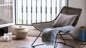 A wicker chair with a cushion and a throw blanket, a woven basket, and a vase with candles on a wooden floor.