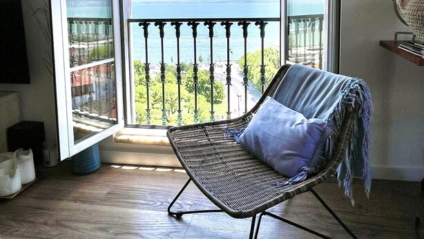 A room with a balcony, a wicker chair with a cushion, a small shelf, and a television on the wall.